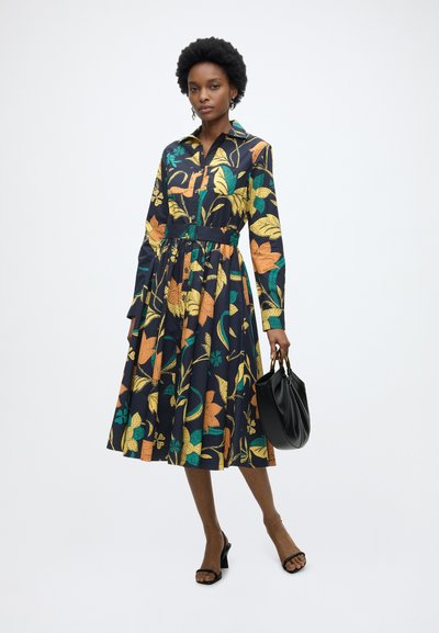 Femme portant une robe ceinturée à manches longues, longueur genoux, avec un motif floral coloré, tenant un sac à main noir et portant des talons noirs.