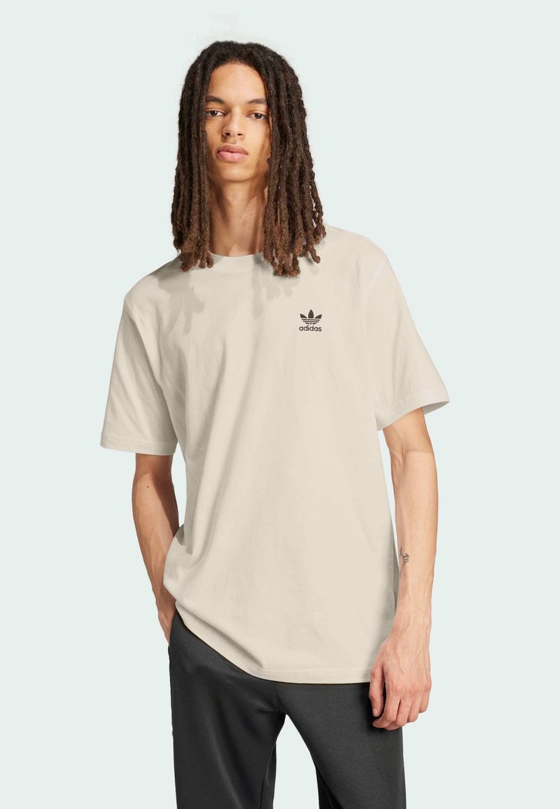 Persona joven con largas rastas, vistiendo una camiseta beige de Adidas y pantalones oscuros, de pie con una mano en el bolsillo.