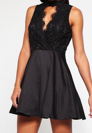 Robe noire sans manches avec corsage en dentelle, décolleté en V profond, taille cintrée et jupe en satin évasée portée par une personne debout devant un fond uni.