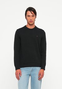 Joven vestido con un suéter negro de cuello redondo con un pequeño logo y jeans azul claro, de pie frente a un fondo blanco liso.