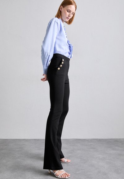 Black flared trousers with side buttons, paired with a light blue blouse. Smooth fabric, structured fit, and decorative button accents.