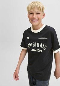 Camiseta negra de manga corta con cuello redondo y borde de las mangas en blanco, que presenta el texto "ORIGINALS studio" y un detalle gráfico de 1990.