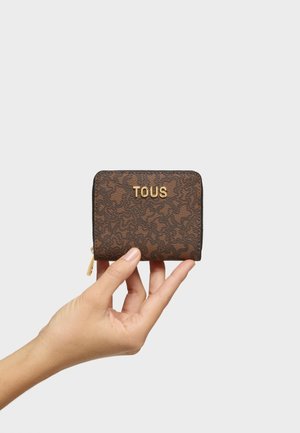 Mano tiene piccolo portafoglio marrone con zip, motivo floreale scuro e logo TOUS dorato sul davanti su uno sfondo chiaro uniforme.