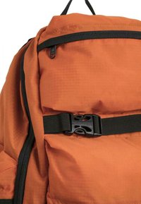 Orange ryggsekk med teksturert stoff, svart glidelås, og justerbar svart stropp med plastspenne for sikker lukking.