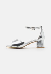 Sølv metallisk ankelsandal med blokkheel. Åpen tå-design, glatt tekstur og en sikker spenne aksent.