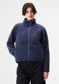 Marineblaue Fleecejacke mit hohem Kragen, Reißverschluss, zwei Fronttaschen und kontrastierenden blauen Reißverschlüssen. Strukturierte, weiche Außenseite.