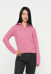 Rosa strikket genser med glidelåskrage, med et teksturert mønster og ribbede mansjetter og hem. Kombinert med mørkegrå bukser.