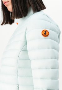 Vrouw in een lichtblauwe gewatteerde jas met een oranje "Save the Duck" logo patch op de bovenarm, gefotografeerd vanuit de zij. 