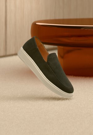Chaussure en daim vert foncé sans lacets avec une semelle blanche, flottant au-dessus d’un sol clair recouvert de moquette et d’une table ronde orange en arrière-plan.