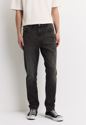 Man die vervaagde zwarte spijkerbroek, wit T-shirt met korte mouwen en zwarte lage sneakers draagt, staande tegen een witte achtergrond.