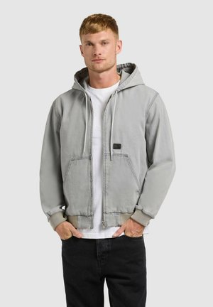 Young man wearing a light gray zip-up hoodie over a white t-shirt and black pants, standing with hands in pockets against plain background.