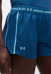 Μπλε αθλητικό σορτς Under Armour με ανοιχτό μπλε περίγραμμα και λογότυπο, σε ψηλό μηρό ενός ατόμου, με το χέρι να φαίνεται στο πλάι.