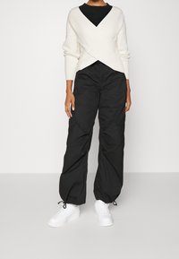 Vit ribbad crossover-tröja tillsammans med svarta lösa byxor. Vita sneakers fulländar outfiten. Enkelt, modernt design med avslappnad passform.