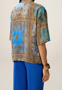 Bluzka z krótkim rękawem w kolorze niebieskim z brązowym, ozdobnym wzorem, z kołnierzykiem i luźnym krojem. Tkanina wydaje się lekka i teksturowana.