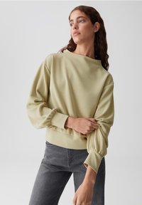 Felpa verde chiaro a maniche lunghe con vestibilità rilassata, caratterizzata da un ampio scollo e polsini arricciati. Abbinata a jeans grigio scuro.