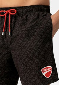 Pantaloni corti neri con motivo ripetuto della scritta "Ducati", vita elastica e cordino rosso. Presenta un logo a scudo rosso con lettering bianco.