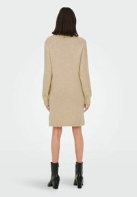 Femme se tenant de dos, vêtue d'une robe en tricot beige à manches longues et de bottines à talons noires sur un fond blanc uni.