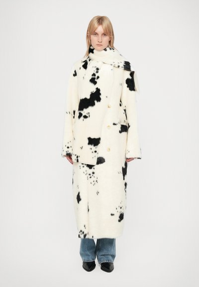 Double-breasted coat made of soft, white fabric with black cow print, featuring two gold buttons and a matching scarf. Worn over blue jeans.