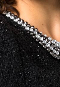 Tissu noir texturé avec une finition scintillante, présentant un col orné de motifs alternant entre des embellissements en cristal noirs et transparents.