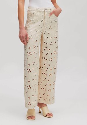 Frau trägt cremefarbene, weit geschnittene Hose mit hohem Bund und floralen Ausschnittmustern und beigefarbene offene, hochhackige Mules, steht mit einer Hand in der Tasche.