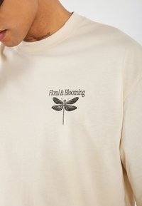Béžové bavlněné tričko s černým potiskem vážky a textem "Floral & Blooming" na levé části hrudi. Krátké rukávy.