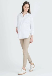 Witte blouse met lange mouwen en knoopsluiting aan de voorkant, met een afgeronde onderkant, gecombineerd met beige strakke broek en witte sneakers met oranje accenten.