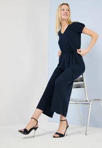 Marinefarbener Jumpsuit mit V-Ausschnitt und kurzen Puffärmelchen, mit gebundenem Taillenbund und cropped Hosenbeinen, kombiniert mit schwarzen Stiletto-Sandalen.