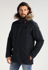 Námořnický modrý parkas s umělou kožešinou na kapuci, s červenými akcenty, zipovými kapsami a voděodolným designem, předvedený na osobě na neutrálním pozadí.
