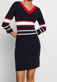 Sukienka do kolan w kolorze granatowym z dekoltem w kształcie litery V oraz czerwonym kołnierzykiem. Posiada poziome pasy w kolorach biało-czerwonych na korpusie i rękawach.