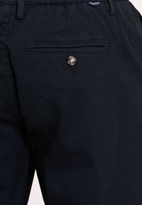 Granatowe spodnie o gładkiej fakturze, z elastycznym pasem i tylną kieszenią zapinaną na guzik. Widoczny subtelny detal w postaci szwów.