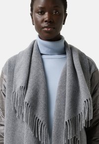 Šedý vlněný šál s třásněmi draped kolem krku, spárovaný s světle modrým rolákem. Měkká textura a útulný vzhled.