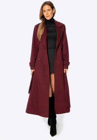 Abrigo largo de mezcla de lana color burdeos con diseño de doble botonadura, solapas con muesca y bolsillos laterales, combinado con un suéter negro de cuello alto y una falda.