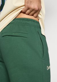 Calças de moletom verdes com uma textura suave, apresentando um bolso traseiro e um pequeno logotipo bordado em creme. Vista parcial com uma camisa de tom claro.