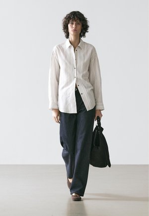 Personne marchant vers l'avant vêtue d'une chemise blanche rayée ample, d'un pantalon large sombre, de chaussures marron, et portant un sac à main noir dans la main droite.