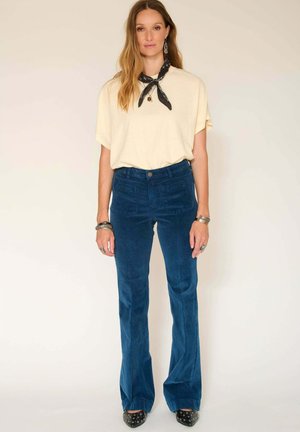 Pantalons en velours côtelé bleu évasés avec poches latérales, associés à un haut à manches courtes ivoire. Les accessoires comprennent une écharpe noire à motifs et des bracelets en argent.