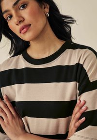 Pull rayé noir et beige avec un col rond. Tissu doux, coupe décontractée, et détails de couture visibles. La main du modèle repose sur le tissu.