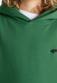 Groene hoodie gemaakt van zachte katoenen stof. Het heeft een capuchon met trekkoord en een klein zwart logo op de borst. Gladde textuur en casual ontwerp.