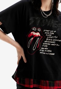 Camiseta de algodón negra con el logo de los Rolling Stones en rojo y blanco, con texto impreso en blanco. Acentuada con strass. Superpuesta sobre una parte inferior de cuadros.