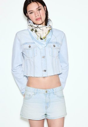 Mladá žena oblečená v krátkej svetlej rifľovej bunde, zodpovedajúcich rifľových kraťasoch a vzorovanej hodvábnej šatke okolo krku, stojaca pred bielym pozadím.