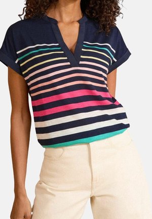 Mujer con una blusa de manga corta azul marino con rayas horizontales multicolores y pantalones beige de talle alto, de pie con una mano en la cadera.