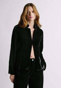 Pikade blondide juustega naine, kes kannab osaliselt avatud mustast sametist bleiserit ja sobivaid pükse, taustal hele ühtlane sein.