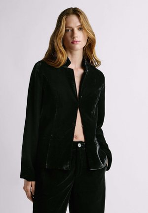 Donna con lunghi capelli biondi che indossa un blazer di velluto nero parzialmente aperto e pantaloni abbinati contro uno sfondo chiaro uniforme.