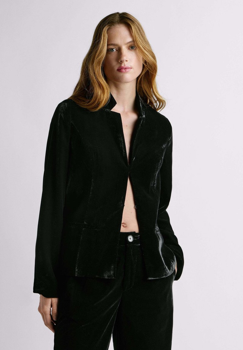 Pikade blondide juustega naine, kes kannab osaliselt avatud mustast sametist bleiserit ja sobivaid pükse, taustal hele ühtlane sein.