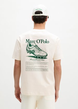 Άτομο που φοράει άσπρο καπέλο και μπλουζάκι Marc O’Polo με μεγάλη πράσινη vintage απεικόνιση ποδοσφαιρικού παπουτσιού και κείμενο στην πλάτη.