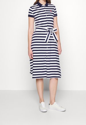 Femme portant une robe polo à rayures horizontales bleu marine et blanches avec des boutons, une ceinture en tissu nouée, et des baskets blanches, debout devant un fond uni.