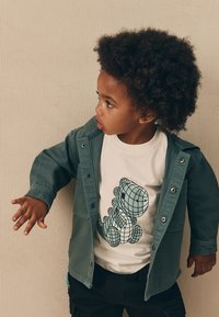 Copil purtând o jachetă verde peste un tricou alb cu grafică cu dinozaur în model grilă. Pantaloni scurți negri, păr creț și fundal neutru.