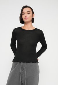 Γυναίκα με κοντά σκούρα μαλλιά, φορώντας μαύρο μακρυμάνικο τοπ και χαλαρά γκρι τζιν, με τα χέρια πίσω από την πλάτη της, στέκεται μπροστά σε ένα απλό φόντο.