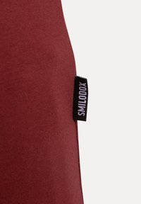 Canotta bordeaux senza maniche realizzata in materiale morbido, con un'etichetta laterale su sfondo nero con testo bianco che riporta il nome del marchio.