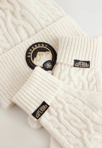 Zestaw kremowej dzianinowej czapki i rękawiczek bez palców, z prążkowaną teksturą i wzorami warkoczowymi. Czarna metka "Superdry" na obu elementach.