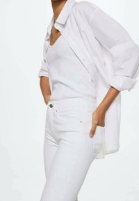 Camisa blanca abotonada sobre una camiseta blanca sin mangas, combinada con vaqueros blancos. El conjunto presenta un ajuste relajado y una tela lisa y texturizada.